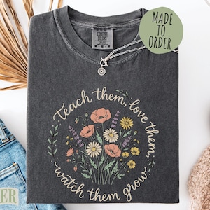 May include: Dark gray Comfort Colors t-shirt with a floral design and the words "Teach them, love them, watch them grow." A silver necklace and a "Made to Order" sticker are visible.