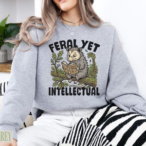 Gray sweatshirt with opossum reading book illustration and "Feral Yet Intellectual" text.