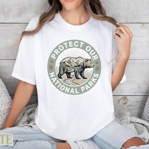 White t-shirt featuring a circular graphic with a bear, mountains, and the text "PROTECT OUR NATIONAL PARKS".