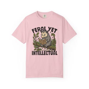 May include: A light pink short-sleeve t-shirt featuring a graphic of an opossum wearing round glasses and reading a book titled Tales & Trivia while perched on a tree branch. The text FERAL YET INTELLECTUAL is printed in black above and below.