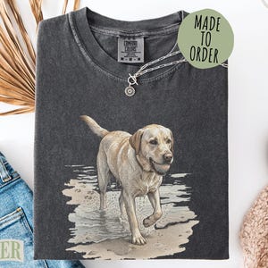 May include: A charcoal gray crew neck t-shirt featuring a graphic illustration of a yellow Labrador retriever standing in shallow water with a ball in its mouth. The shirt has a Comfort Colors label at the neck and a silver toggle necklace.