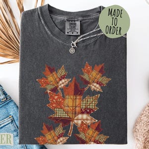 May include: A charcoal gray short-sleeve t-shirt featuring a graphic print of five maple leaves designed with a patchwork pattern of various autumn-colored fabrics including plaid, floral, and solid orange, red, and brown tones.