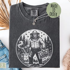 May include: Dark gray t-shirt with a graphic of a Bigfoot beekeeper holding a smoker and a honeycomb frame, surrounded by bees and trees. The shirt has the text "MADE TO ORDER" on a green circle.