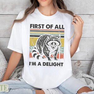 White t-shirt with raccoon graphic and text "First of all I'm a delight".