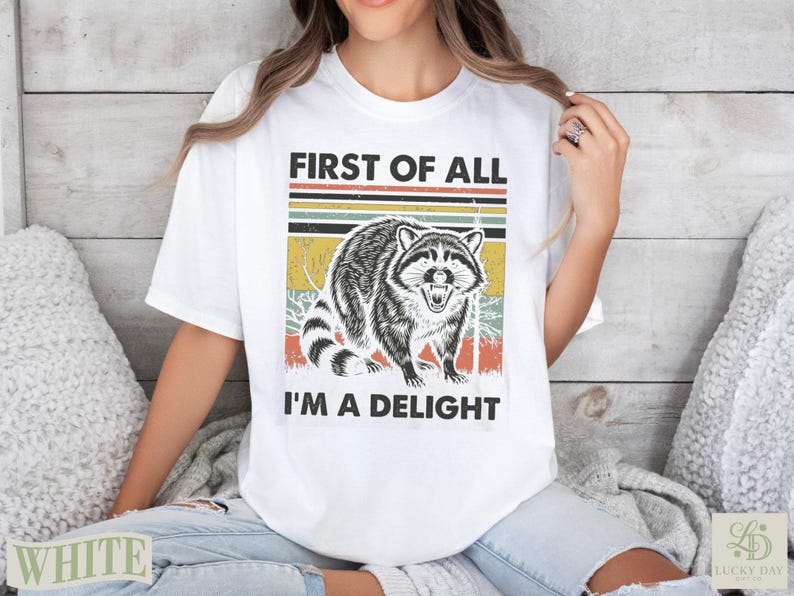 White t-shirt with raccoon graphic and text "First of all I'm a delight".