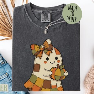 May include: A charcoal gray t-shirt featuring a graphic of a cream-colored ghost with a patchwork pattern, wearing a large plaid bow and holding a small pumpkin. The shirt has a crew neckline and a Comfort Colors brand label at the inner collar.
