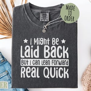 May include: Dark gray t-shirt with the text "I Might Be Laid Back But I Can Lean Forward Real Quick" in white. The shirt has a crew neck and is folded. A silver necklace and a "Made to Order" sticker are visible.