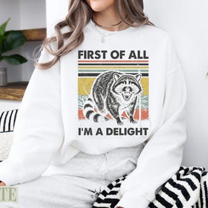 White sweatshirt with a raccoon graphic and text "FIRST OF ALL I'M A DELIGHT".