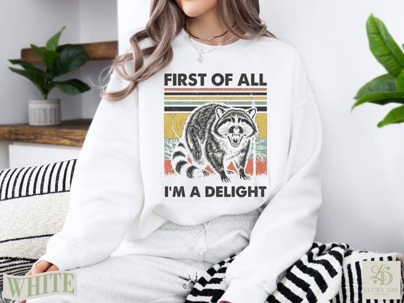 White sweatshirt with a raccoon graphic and text "FIRST OF ALL I'M A DELIGHT".