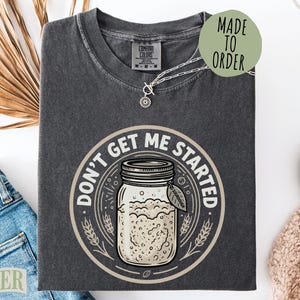 May include: Dark gray Comfort Colors t-shirt with a circular graphic that says "DON'T GET ME STARTED" around a mason jar filled with a grainy substance. The shirt has a crew neck and short sleeves. A silver necklace with a small pendant is visible.