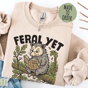 Tan sweatshirt with opossum reading "Feral Yet Tales & Trivia."