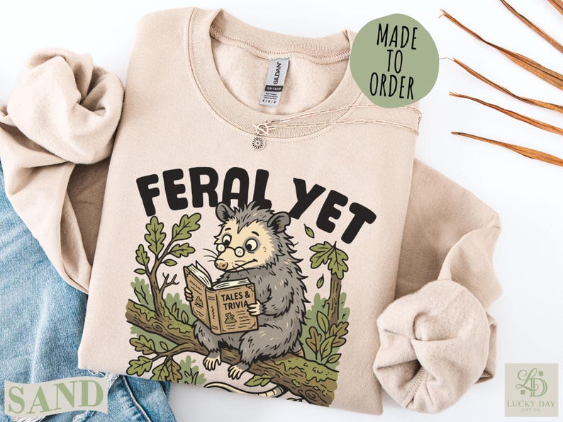 Tan sweatshirt with opossum reading "Feral Yet Tales & Trivia."