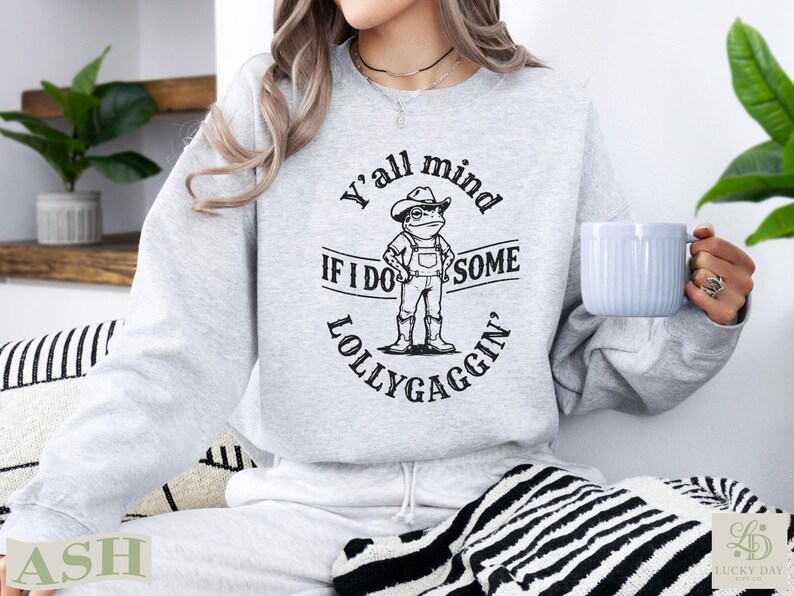Light gray sweatshirt with cartoon frog wearing a cowboy hat and bib overalls, text reads "Y'all mind if I do some lollygaggin'".