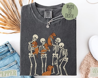 May include: A charcoal gray short-sleeve t-shirt featuring a graphic print of four human skeletons interacting with five brown dachshund dogs. The shirt has a crew neckline and a Comfort Colors brand label at the inner neck. A green circular tag reads MADE TO ORDER.