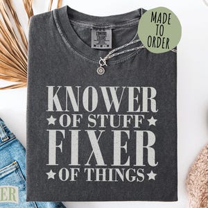 May include: Dark gray t-shirt with the text "KNOWER OF STUFF FIXER OF THINGS" in white. The shirt is folded and has a small silver necklace. A green circle with "MADE TO ORDER" is on the shirt.