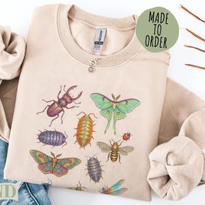 Beige sweatshirt with insects: moth, beetle, dragonfly, bee, roly-poly, and butterfly.