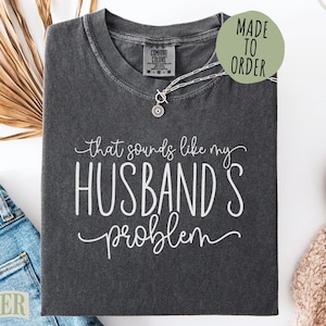 May include: Dark gray t-shirt with the phrase "that sounds like my husband's problem" in white script. The shirt has a crew neck and is folded. A silver necklace with a pendant is on the shirt. A "Made to Order" sticker is on the shirt.