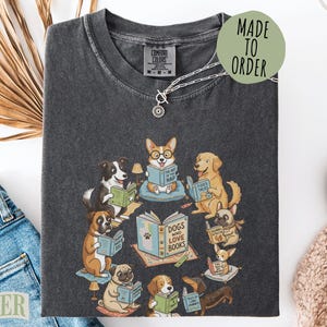 May include: A charcoal gray t-shirt featuring a graphic illustration of various dog breeds sitting in a circle and reading books. The central book is titled Dogs Who Love Books, surrounded by dogs with individual books and small lamps.
