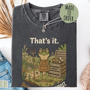 May include: Dark gray t-shirt with a frog graphic holding a pitchfork, standing in a garden scene with the text "That's it. You're compost." A "Made to Order" sticker is on the top right.