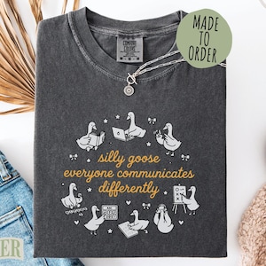 May include: Dark gray Comfort Colors t-shirt with a whimsical design featuring cartoon geese interacting with books, laptops, and other items. The text reads "silly goose everyone communicates differently". A silver necklace is on the shirt.