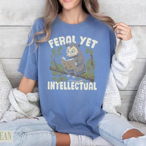 Blue t-shirt with opossum reading a book and text that says "Feral Yet Intellectual".