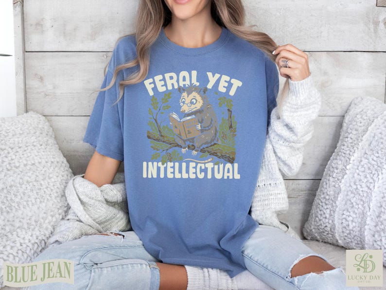 Blue t-shirt with opossum reading a book and text that says "Feral Yet Intellectual".