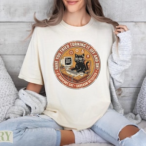 May include: A charcoal gray t-shirt featuring a circular graphic of a black cat wearing a headset and glasses, sitting at a computer desk. The text reads: HAVE YOU TRIED TURNING IT OFF AND ON? IT SUPPORT CAT. SYSTEM ERROR: REBOOT? NOPE.