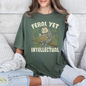 Olive green t-shirt with opossum reading a book, "Feral Yet Intellectual."