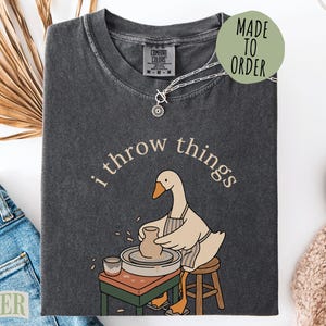 May include: A charcoal gray short-sleeve t-shirt featuring a graphic of a goose wearing an apron and working at a pottery wheel. The text "i throw things" is printed in a light beige arc above the goose. The shirt has a crew neckline.