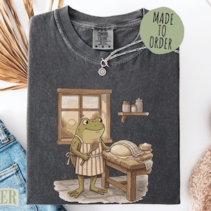 May include: A charcoal gray t-shirt featuring a graphic of a green frog wearing a striped apron, standing at a wooden table with a loaf of dough. The frog holds a wooden spoon. A window, a shelf with jars, and the text MADE TO ORDER are visible.