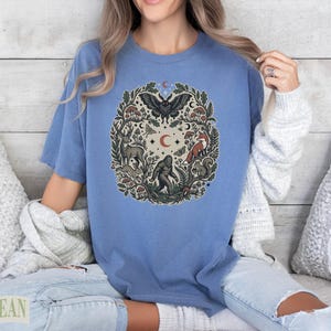 Blue t-shirt featuring forest animals, mushrooms, and celestial motifs in a circular design.