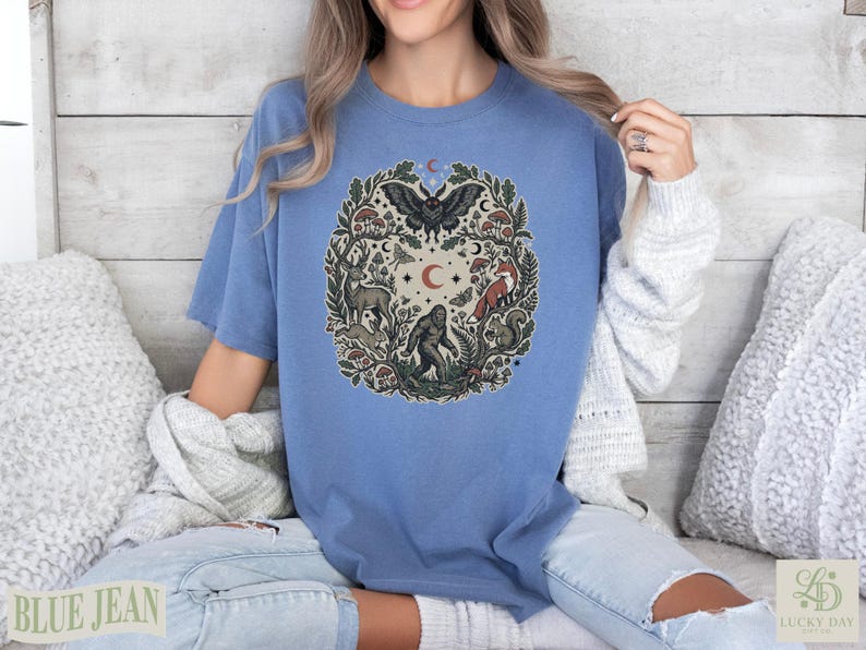 Blue t-shirt featuring forest animals, mushrooms, and celestial motifs in a circular design.