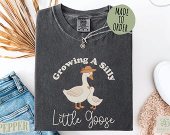 May include: A charcoal gray t-shirt featuring a graphic of an adult goose wearing a tan hat standing next to a small gosling. The text "Growing A Silly Little Goose" is printed in white above and below the geese. The shirt has a Comfort Colors label.