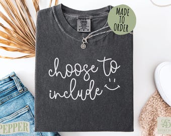 May include: A dark gray t-shirt with the words "choose to include" in white script, along with a smiling face. A silver necklace with a circular pendant rests on the shirt. The shirt is folded on a white surface, with a pair of jeans and a beige sweater nearby.