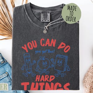 May include: A charcoal gray t-shirt featuring a graphic with the red text "YOU CAN DO HARD THINGS" and blue text "you got this!". The design includes cartoon illustrations of a pencil, an apple, and a book with arms and legs. The shirt has a crew neck.