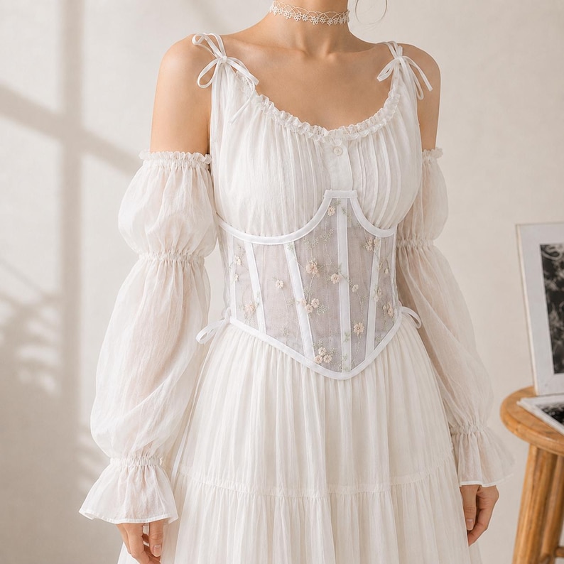May include: A white dress featuring long, sheer, puffed sleeves with ruffled cuffs and shoulder cutouts. The bodice includes a structured, semi-sheer corset overlay with white piping and delicate floral embroidery, tied at the shoulders with thin ribbons.