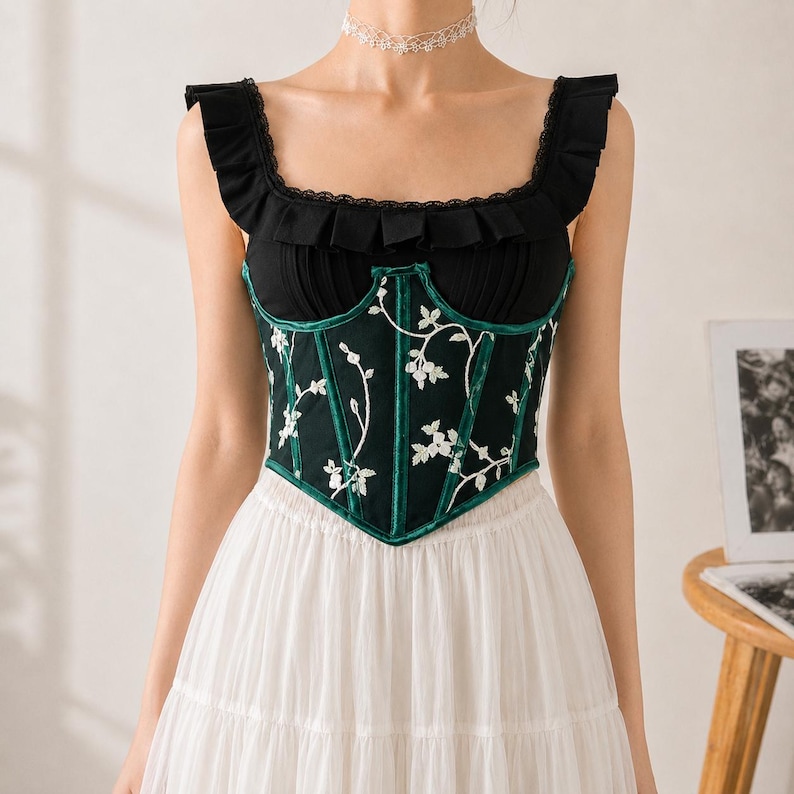 May include: A sleeveless black corset top featuring a square neckline with ruffled trim, a dark green velvet-trimmed bodice with white floral embroidery, and a pointed hemline, paired with a long, layered white skirt and a white lace choker.