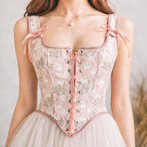 May include: A pink corset with a floral and bunny print. The corset has a square neckline, ribbon straps, and a lace-up front. The edges are trimmed with pink piping, and the straps are tied with bows. The corset is part of a dress.