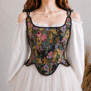 May include: A floral tapestry corset with black trim and shoulder straps. The corset features a vibrant pattern of pink, yellow, and blue flowers on a black background. The straps are black and tied with bows. The corset is worn over a white off-the-shoulder blouse and a white tulle skirt.