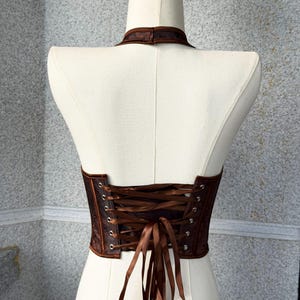 May include: A brown corset with a halter neckline and a lace-up back. The corset is made of a textured fabric and features a satin ribbon for the lacing. The garment is displayed on a white mannequin.