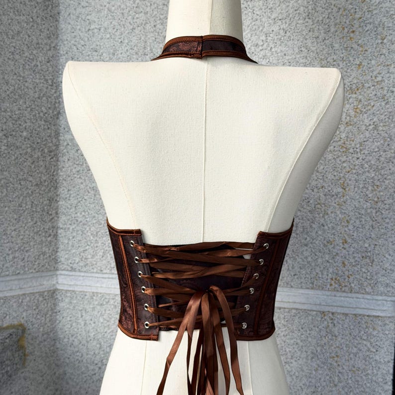 May include: A brown corset with a halter neckline and a lace-up back. The corset is made of a textured fabric and features a satin ribbon for the lacing. The garment is displayed on a white mannequin.