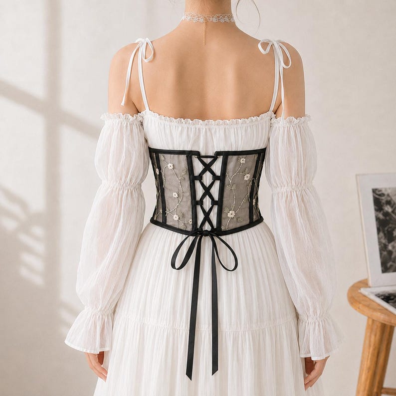 May include: A white long-sleeved dress featuring a sheer black corset-style waist panel with white floral embroidery. The corset has a black lace-up back closure with a ribbon tie. The dress has off-the-shoulder sleeves and thin shoulder straps.