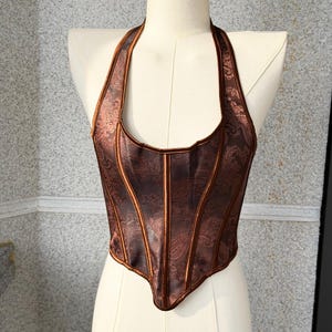 May include: A brown halter top corset with a paisley pattern. The corset has vertical panels and a structured bodice. The halter straps and edges are a darker brown, creating a contrast. The corset is displayed on a mannequin.