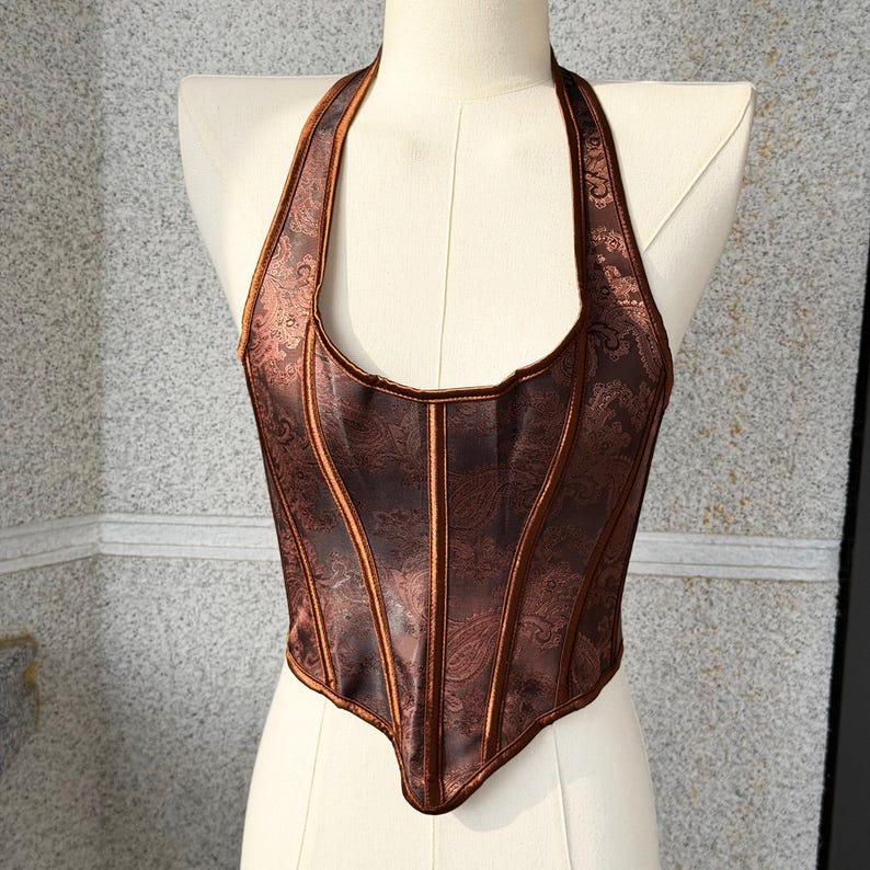 May include: A brown halter top corset with a paisley pattern. The corset has vertical panels and a structured bodice. The halter straps and edges are a darker brown, creating a contrast. The corset is displayed on a mannequin.