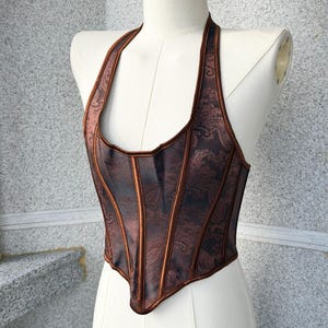 May include: A brown halter-neck corset with a paisley pattern. The corset has vertical panels and a contrasting trim. The garment is displayed on a mannequin against a neutral background. The corset is a dark brown color.