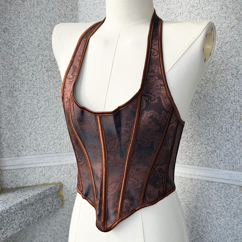 May include: A brown halter-neck corset with a paisley pattern. The corset has vertical panels and a contrasting trim. The garment is displayed on a mannequin against a neutral background. The corset is a dark brown color.