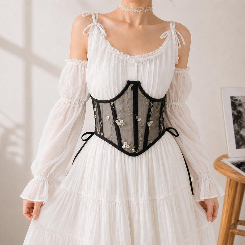 May include: A white dress with long, off-the-shoulder sleeves and a tiered skirt, featuring a black corset-style waist cincher made of sheer mesh fabric with white floral embroidery and black trim, tied at the sides with thin black ribbons.