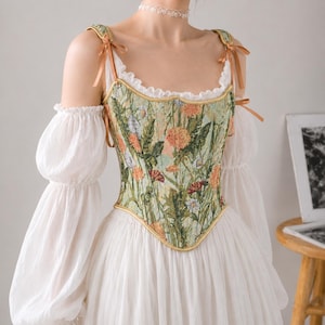 May include: A corset top featuring a green floral tapestry pattern with orange and white flowers, gold trim, and ribbon shoulder ties. It is worn over a white blouse with long, sheer, puffed sleeves and a ruffled neckline, paired with a white skirt.
