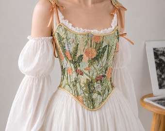 Könnte beinhalten: Ein Korsett-Oberteil mit grünem Blumen-Tapestry-Muster, orangefarbenen und weißen Blüten, goldener Umrandung und Schulterbändern. Es wird über einer weißen Bluse mit langen, transparenten Puffärmeln und einem gerüschten Ausschnitt getragen.