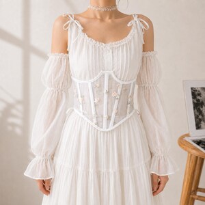 May include: A white dress featuring a sheer corset-style bodice with delicate floral embroidery, long off-the-shoulder sleeves with ruffled cuffs, and shoulder ties. The bodice is structured with vertical white piping and a scalloped hemline.